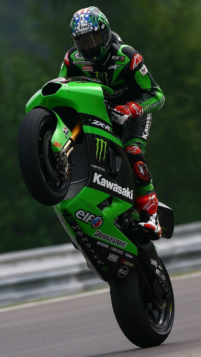 Motorcycle racer performing a wheelie on a green Kawasaki sportbike with a star-spangled helmet on a track.
