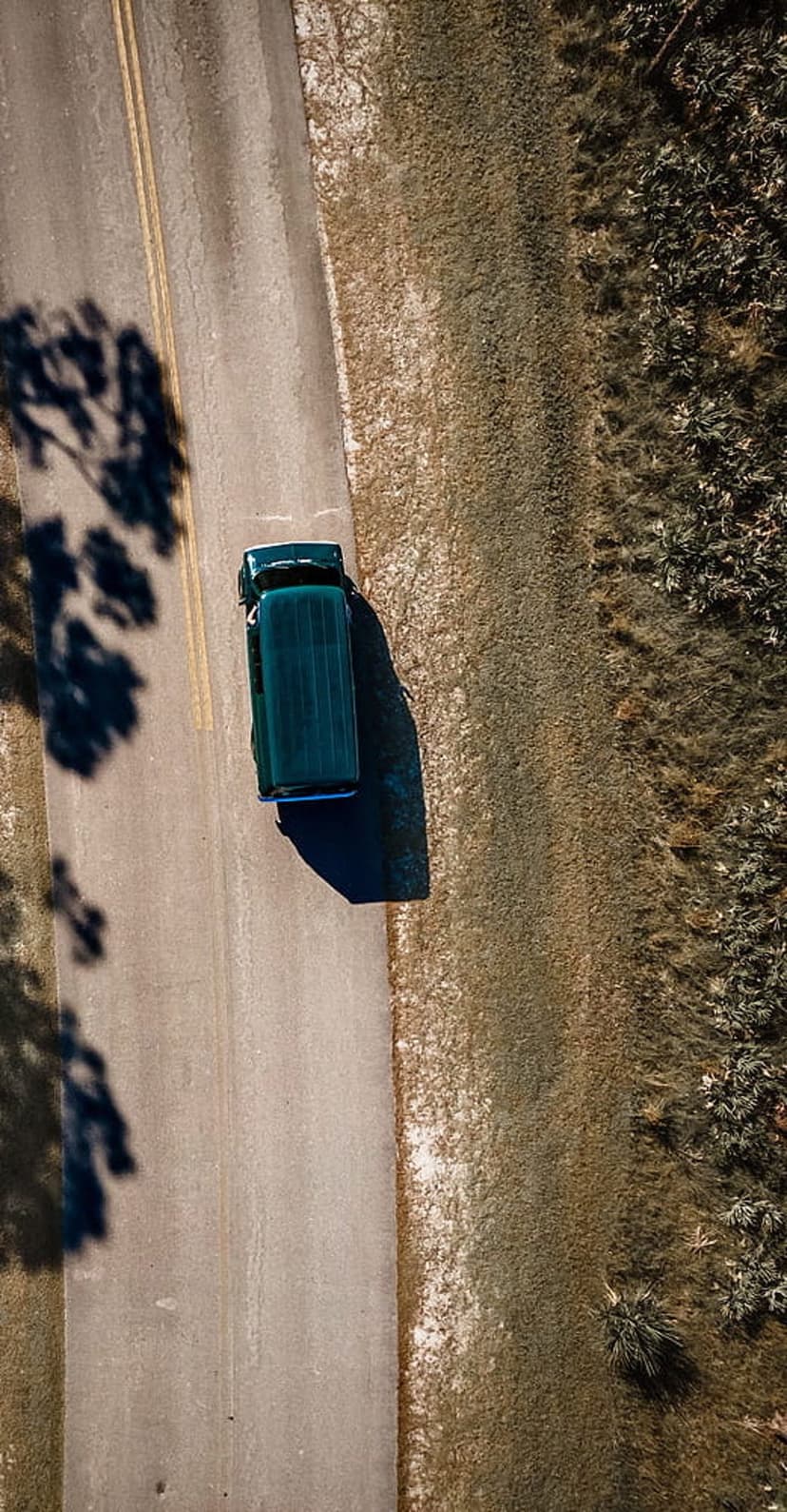 Green van driving on an asphalt road with a yellow center line and long tree shadows across the surface.