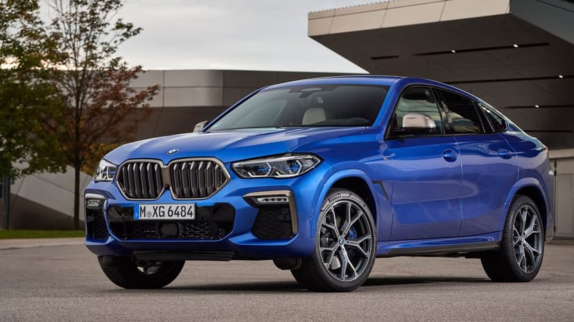 Blue BMW X6 M luxury SUV parked on asphalt in front of a modern building with autumn trees in the background.
