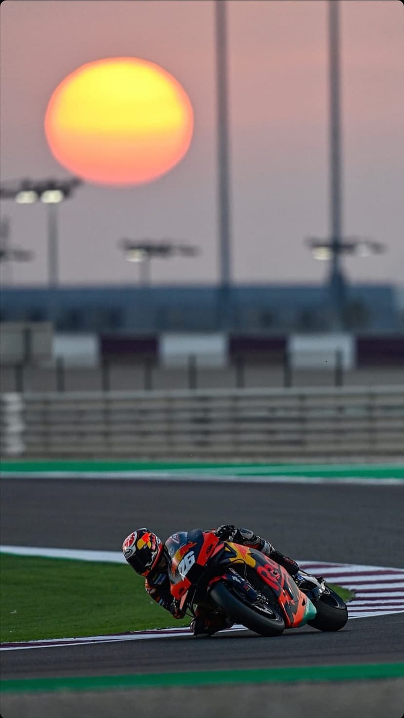 Motorcyclist in a red racing suit leans an orange KTM bike into a sharp turn against a large orange sunset.