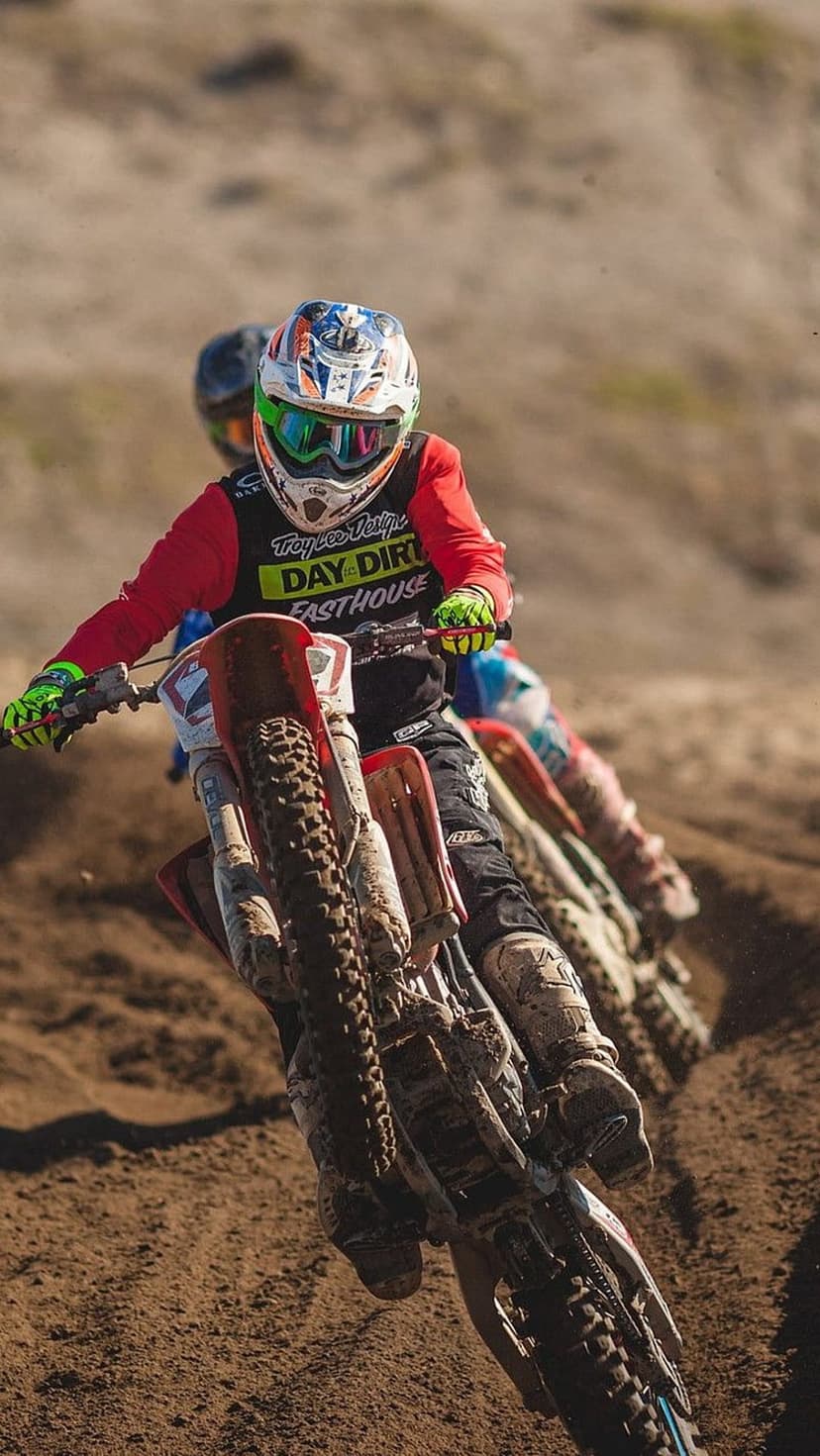 Motocross rider in a red racing suit and helmet navigates a sandy track amid thick clouds of kicked-up dust.