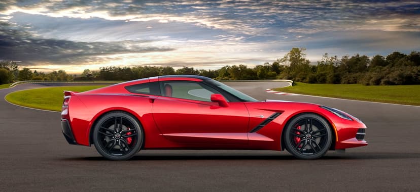 Red Chevrolet Corvette C7 parked on an asphalt racetrack with a dramatic sunset sky and green fields.