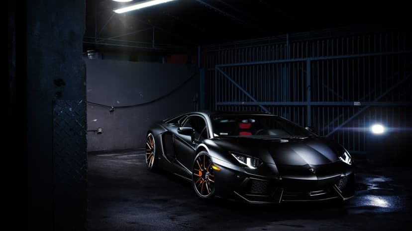 Matte black Lamborghini Aventador with orange wheels parked in a dark industrial garage under dramatic lighting.