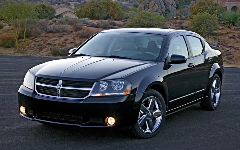Black Dodge Avenger sedan with lit fog lights parked on asphalt against a rocky desert landscape.
