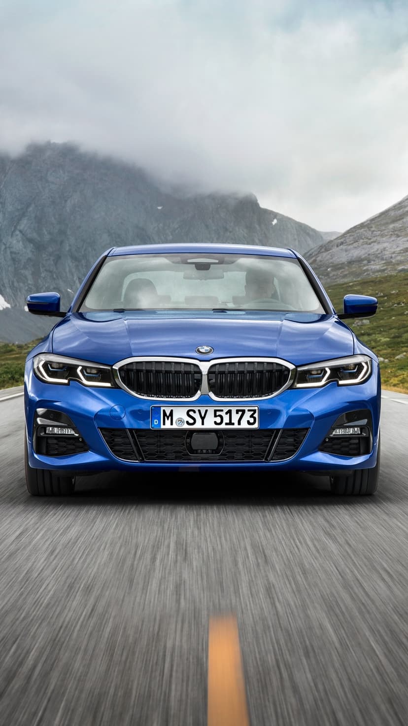 Front view of a blue BMW sedan driving on a mountain road with misty peaks and a cloudy sky background.