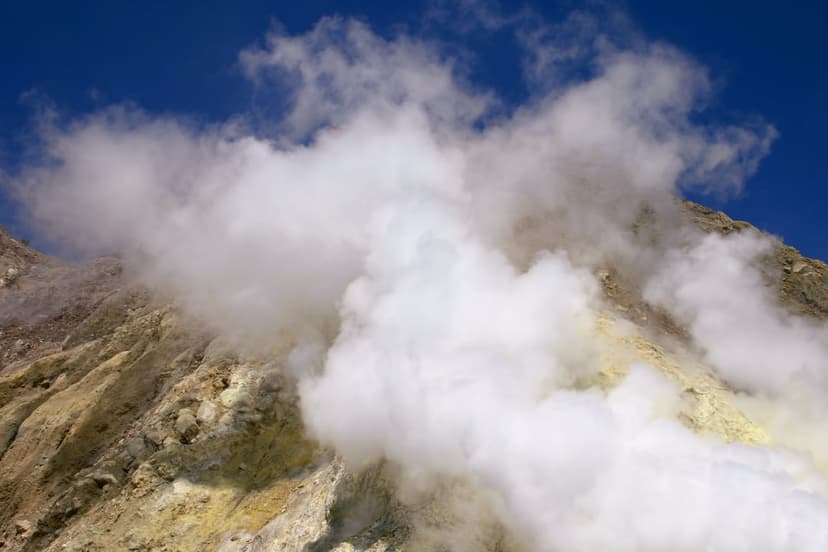 Volcanic crater with rising steam plumes, tan rocky slopes, and yellow sulfur crystals under a clear blue sky.