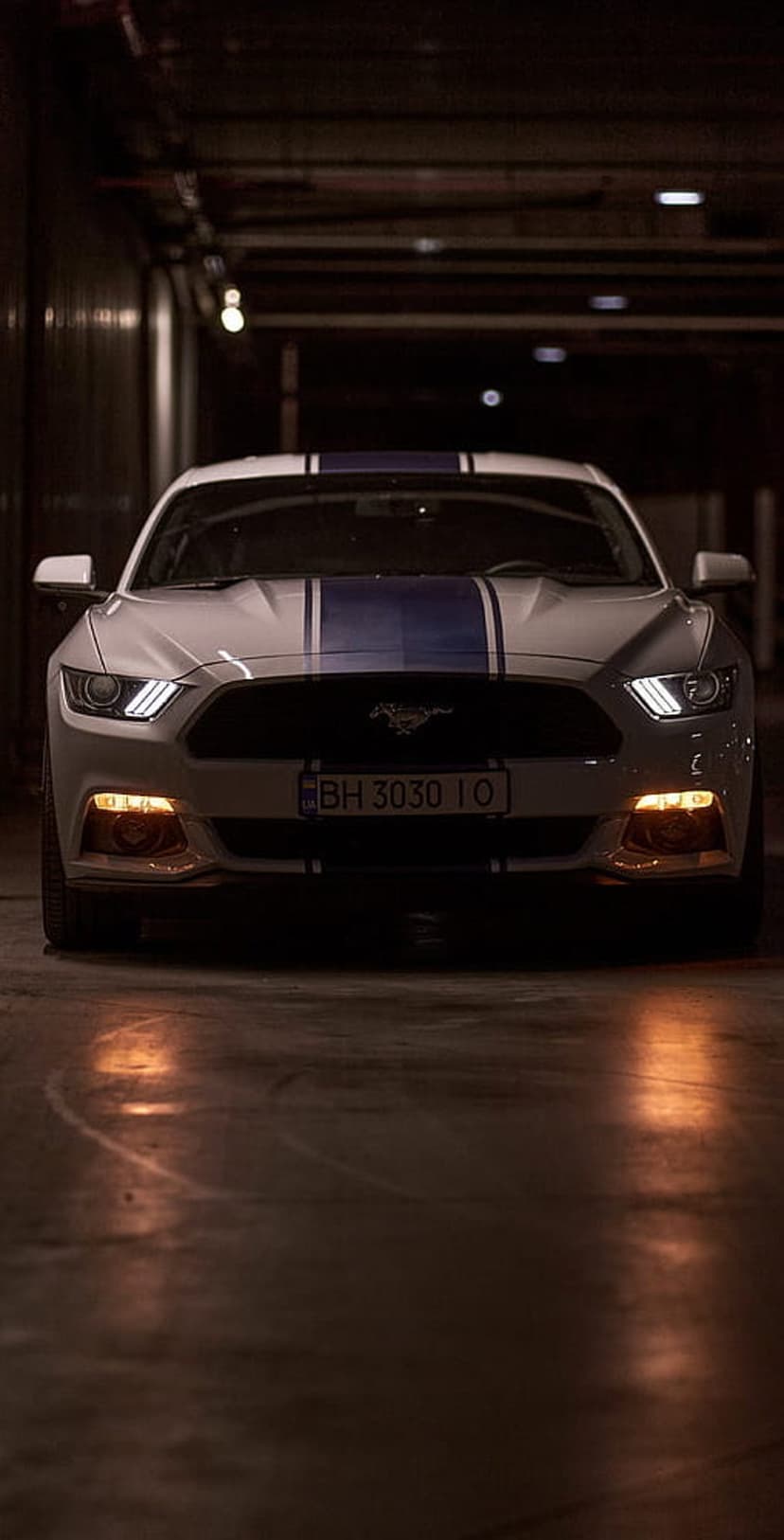 White Ford Mustang GT with blue stripes parked in a dark tunnel with glowing headlights on a concrete floor.