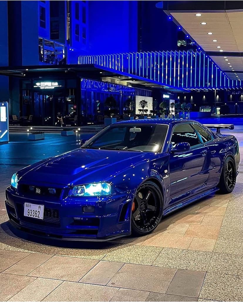 Nissan Skyline R34 GT-R with a blue finish parked before a building with glowing azure architectural lighting.
