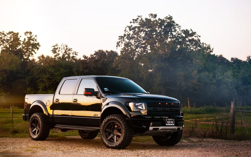 Black Ford F-150 Raptor pickup truck on a gravel road with golden sunset reflections and a forest background.