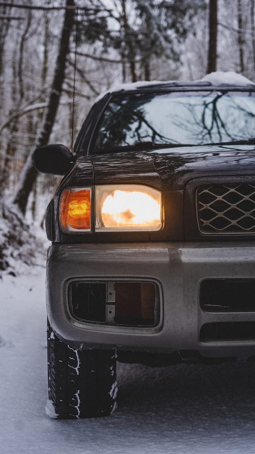 SUV headlight and amber turn signal glow against a snowy forest backdrop with snow-caked tire treads at dusk.