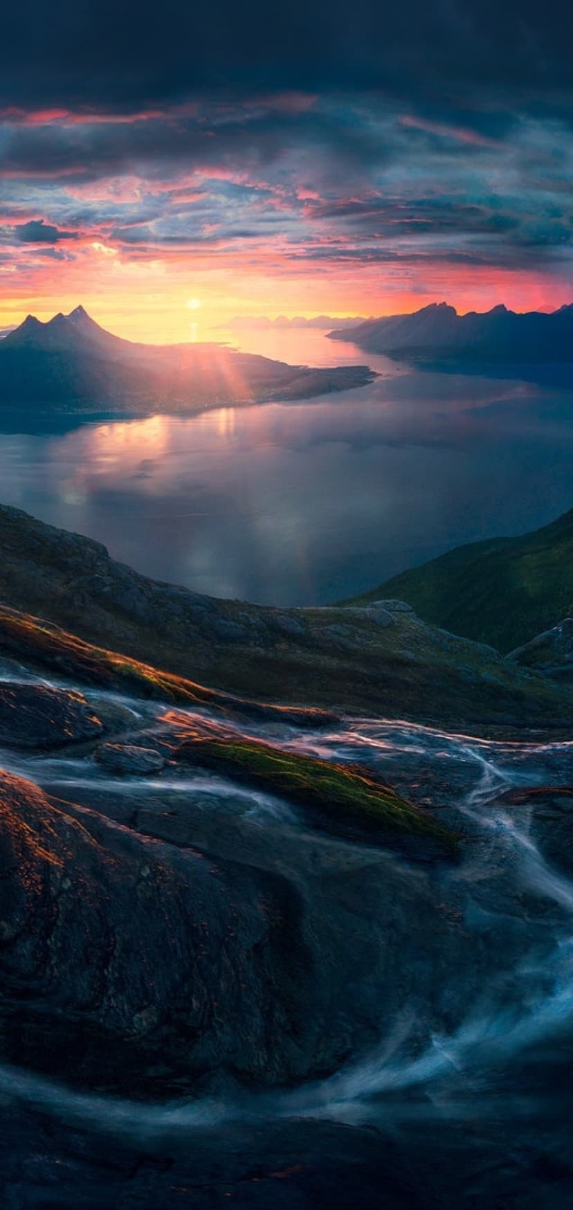 Jagged mountains tower over a calm fjord as a small waterfall flows over rocks beneath a pink sunrise sky.