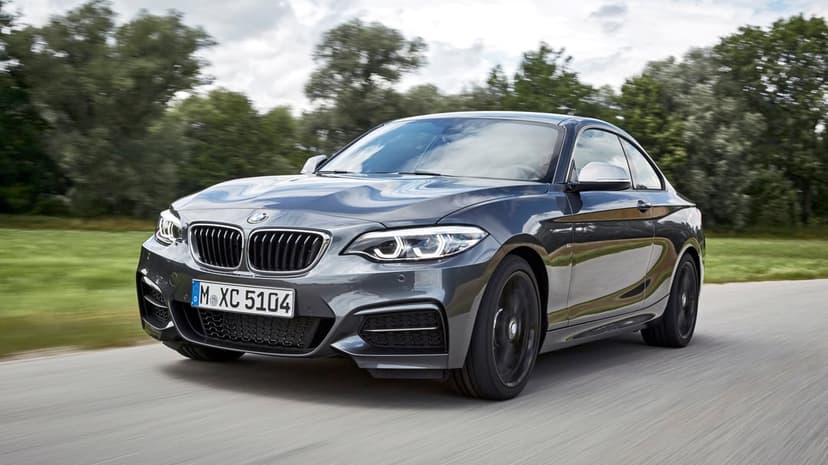Dark grey BMW 2 Series coupe driving on a road with motion blur against a background of green trees.
