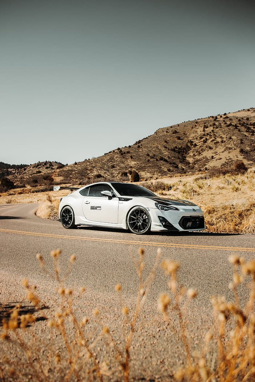 White Toyota GT86 with a black hood cruises on a winding asphalt road through arid, sunlit mountain peaks.