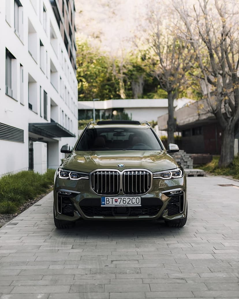 Green BMW X7 SUV with a signature grille parked on a driveway beside a modern white house and green trees.