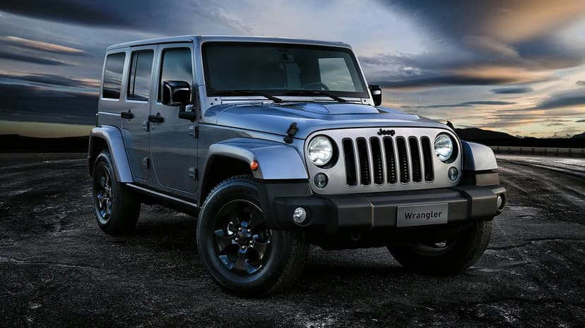 Silver Jeep Wrangler 4x4 SUV with black wheels on gravel at dusk under a cloudy sunset sky with dramatic lighting.