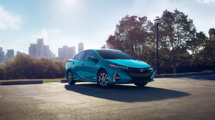 Teal Toyota Prius Prime parked in a lot with a city skyline and trees under a bright blue sunny sky.