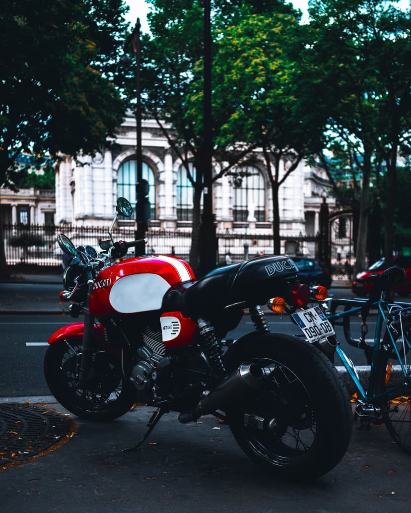 Red Ducati motorcycle with white accents parked on a city street in front of a classical stone building and trees.