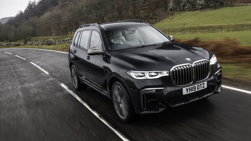 Black BMW X7 driving fast on a wet winding road through green hills under a cloudy sky with motion blur.
