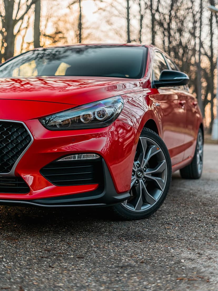 Red Hyundai i30 N Line front grille and headlights reflecting golden sunset light against a blurred forest.