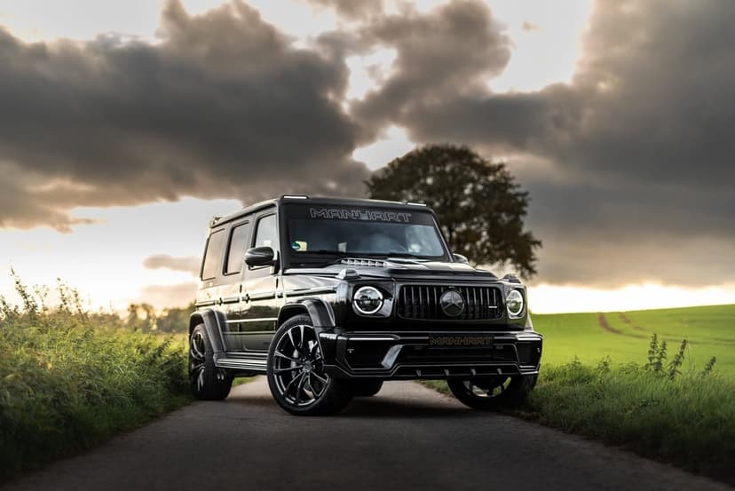 Black Manhart Mercedes-Benz G-Class SUV parked on a narrow road under a dramatic, cloudy sunset sky with green fields.