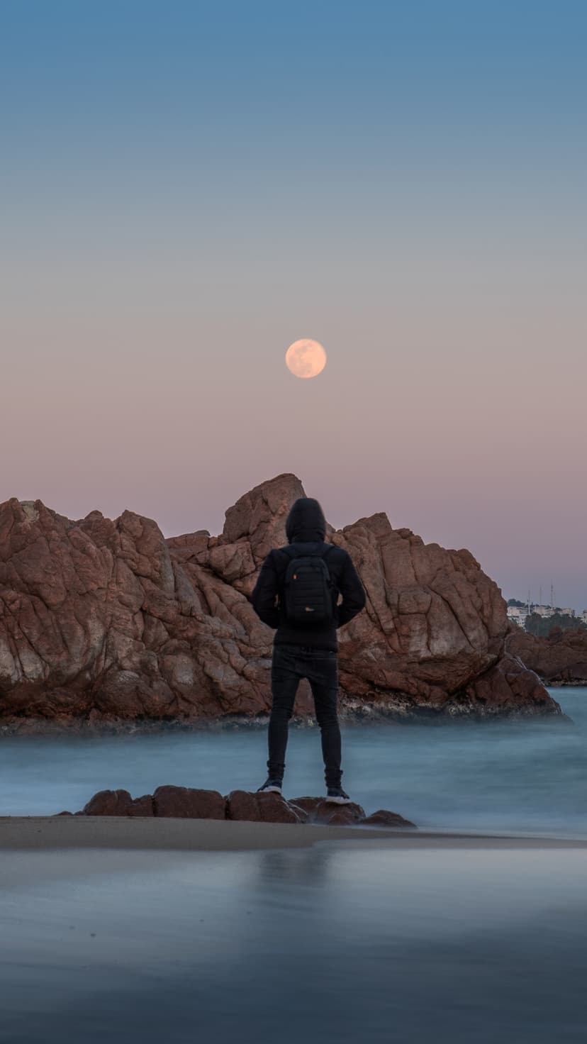 Hooded silhouette standing on coastal rocks facing a large full moon over misty, long-exposure ocean waters.