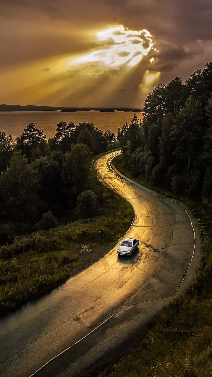 Silver car drives on a wet winding road through a forest as sunbeams pierce through dark clouds over a lake.