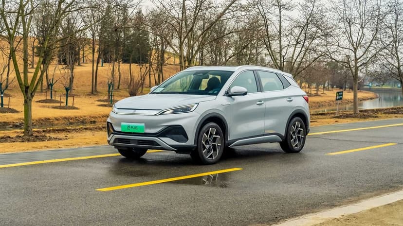 Silver electric SUV parked on a paved road with yellow lines, surrounded by bare winter trees and a park pond.