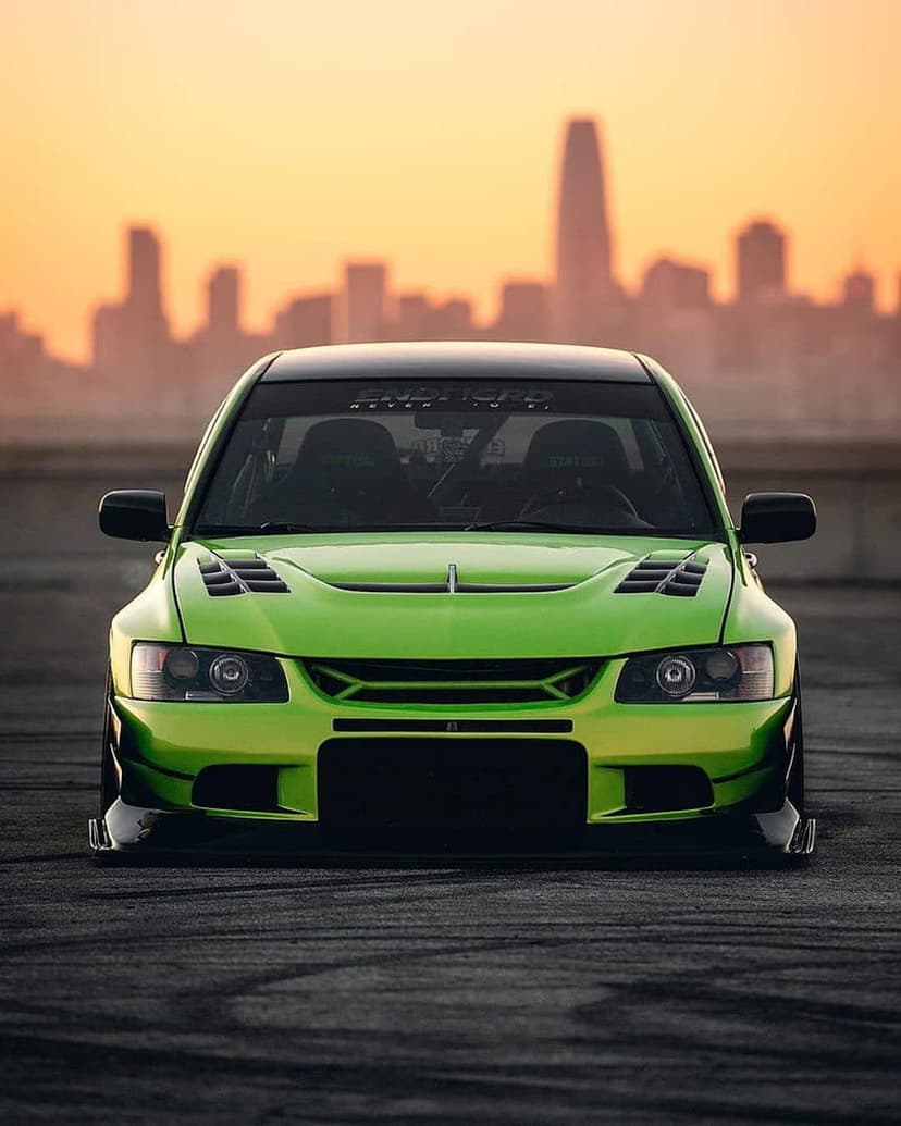 Green Mitsubishi Evolution parked on asphalt with tire marks before a blurred sunset city skyline background.