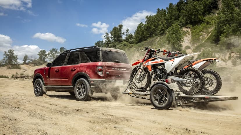 Red Ford Bronco Sport towing two orange dirt bikes through a dust cloud on a rugged dirt road under a clear sky.