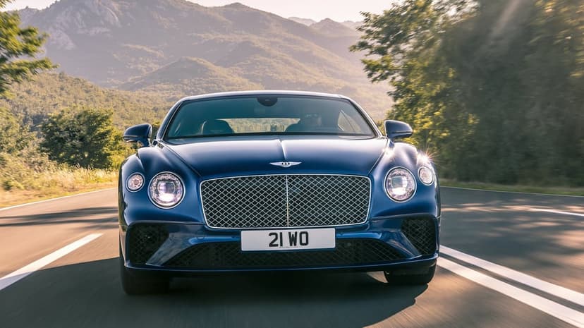 Blue Bentley Continental GT driving on a mountain road with warm sunlight and hazy peaks in the background.