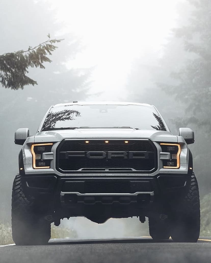 Ford Raptor truck with glowing headlights drives on a misty forest road past tall evergreen trees.