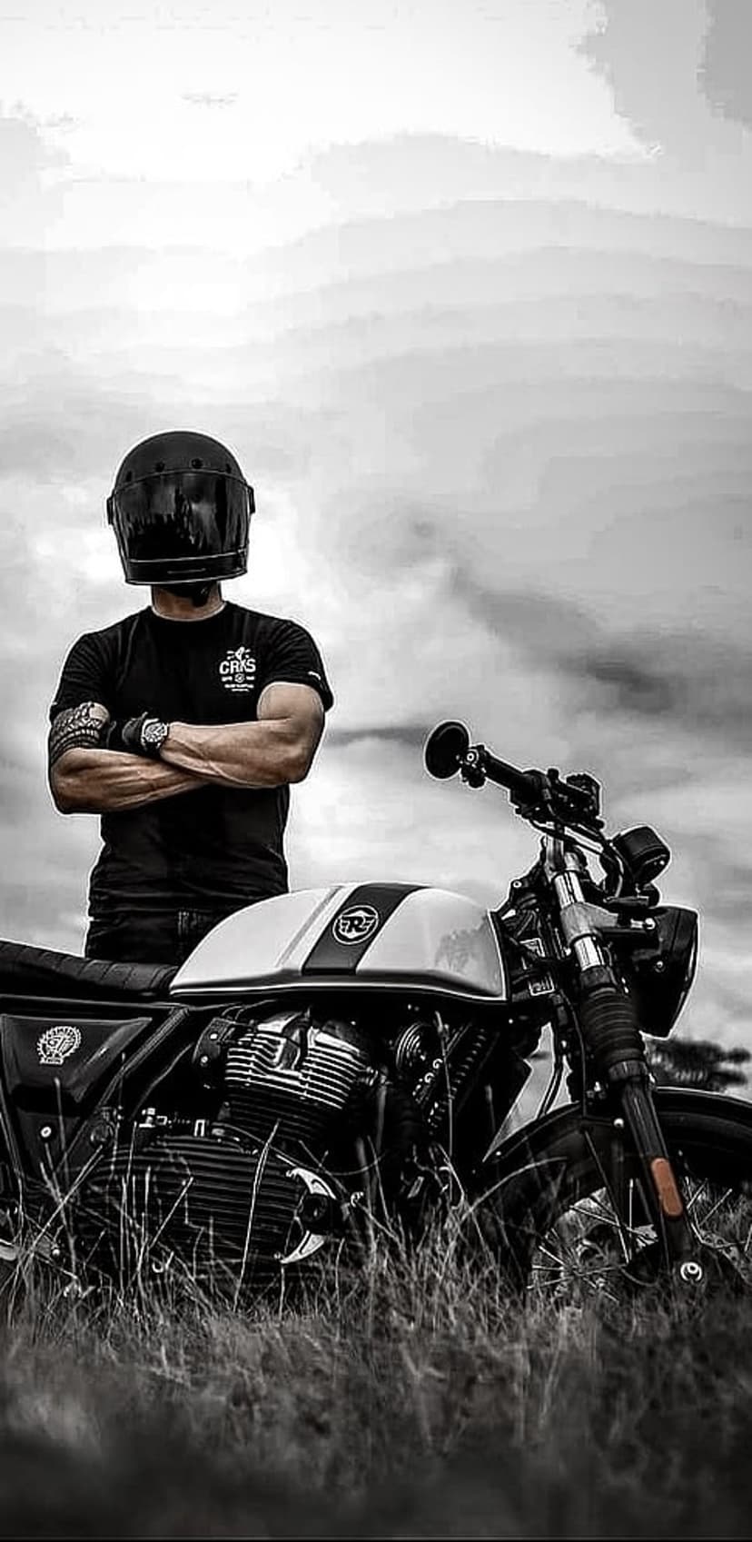 Motorcyclist in a full-face helmet stands by a vintage bike engine under a cloudy sky in a monochrome field.