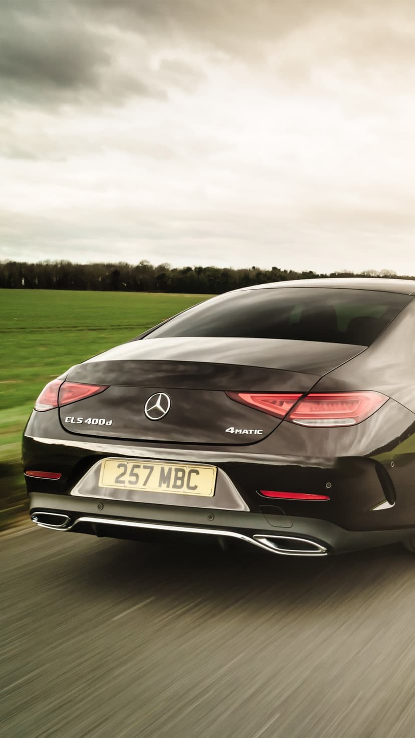 Rear view of a dark Mercedes-Benz CLS 400d driving past a green field under a moody cloudy sunset sky.