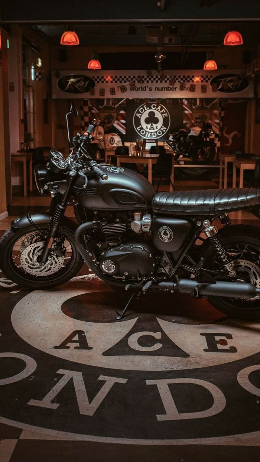 Black vintage motorcycle parked on an Ace Cafe floor logo illuminated by warm orange pendant lights in London.