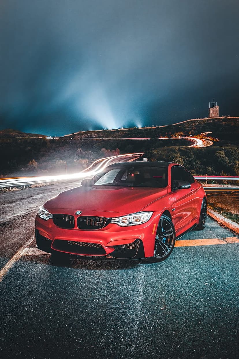 Red BMW M4 parked on a wet road at night with distant light streaks and a starry sky in the background.