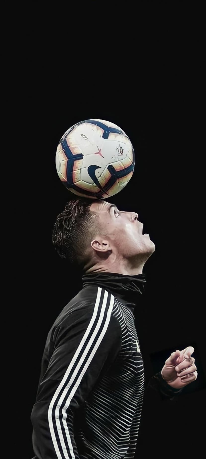 Cristiano Ronaldo balances a white soccer ball on his forehead while wearing a black Adidas tracksuit.