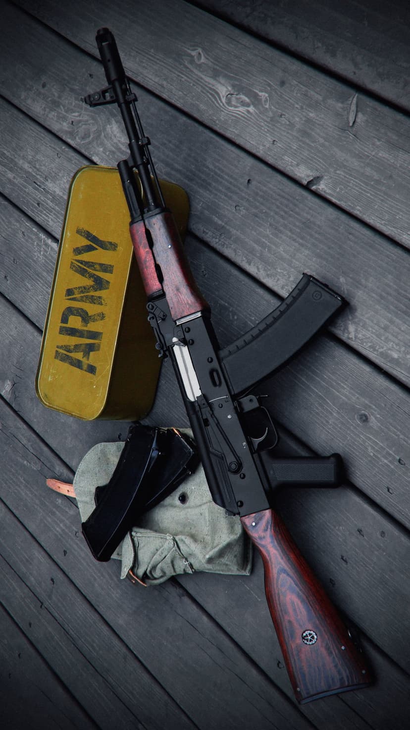 AK-47 rifle with a wooden stock lies next to a yellow ammunition crate and camouflage cap on a dark table.