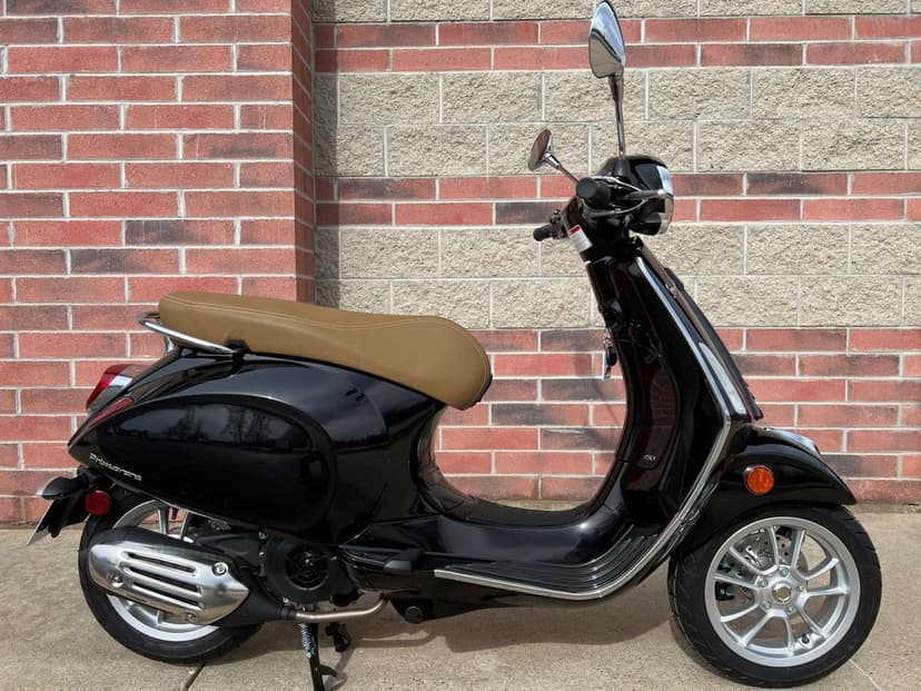 Black Vespa Primavera scooter parked against a brick wall, showing its tan seat and chrome details in daylight.