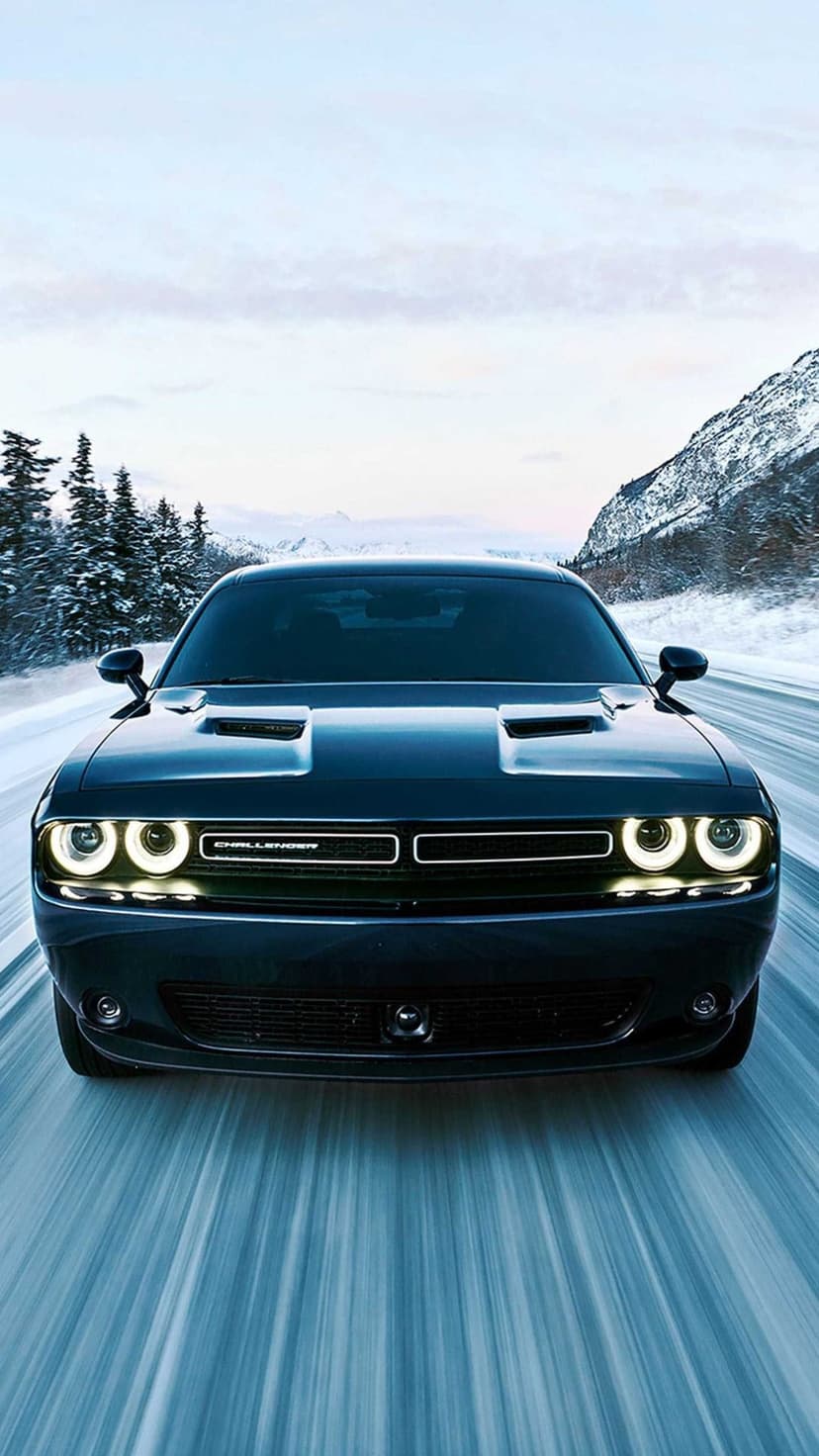 Dark Dodge Challenger with glowing halo headlights driving on a snowy road through pine-covered mountains.
