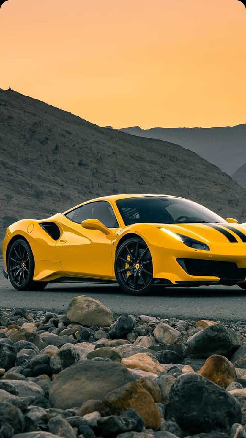 Yellow Ferrari 488 GTB with black stripes parked on a rocky coast beneath an orange mountain sunset sky.