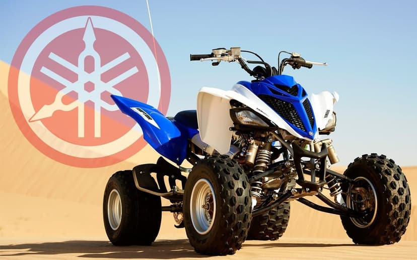 Blue and white Yamaha Raptor ATV on desert sand dunes with a large red Yamaha logo overlay in the sky.