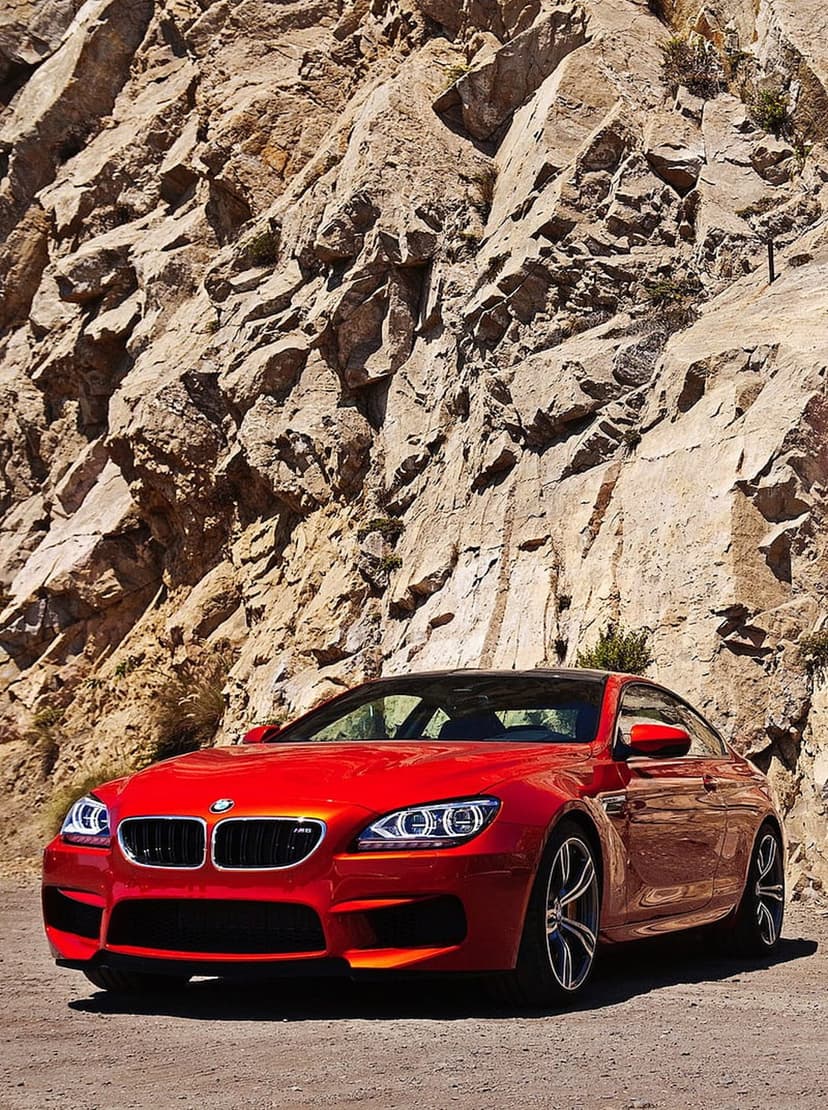 Red BMW M6 Coupe parked on a dusty dirt road in front of a massive textured mountain rock face at a low angle.