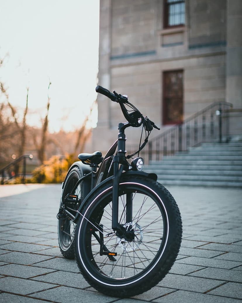 Black electric fat tire bicycle with white-walled tires parked on a stone walkway near urban stairs and trees.