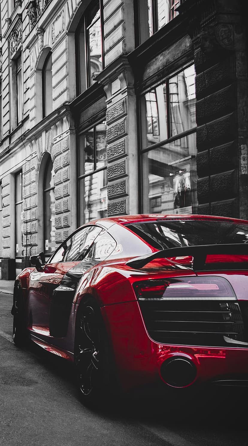 Red Audi R8 supercar parked on a cobblestone street beside ornate European buildings and tall glass windows.