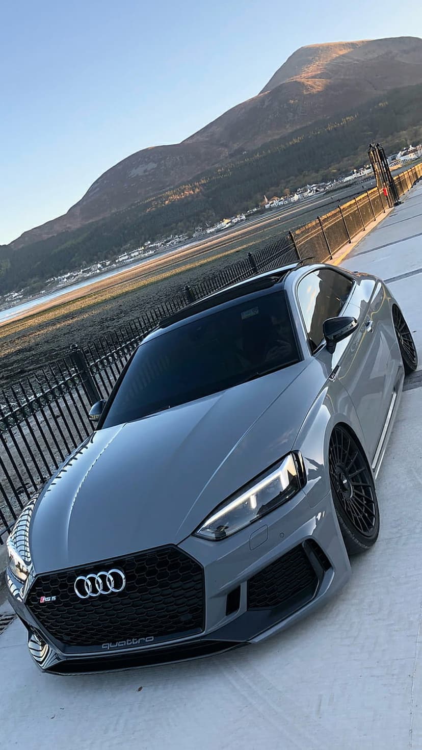 Grey Audi RS5 Coupe with black wheels parked near a mountain lake under a clear blue sky on a promenade.