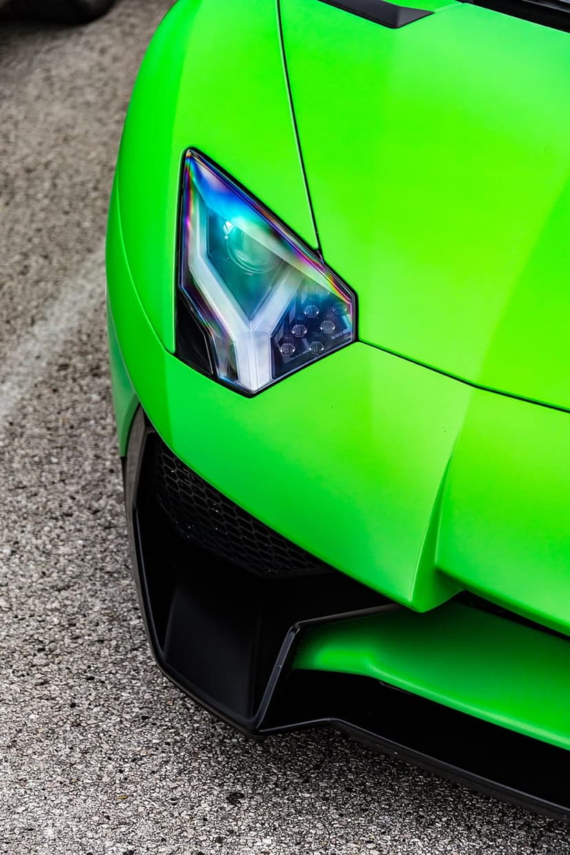 Lamborghini Aventador headlight with LED accents and a green front bumper resting on textured black asphalt.