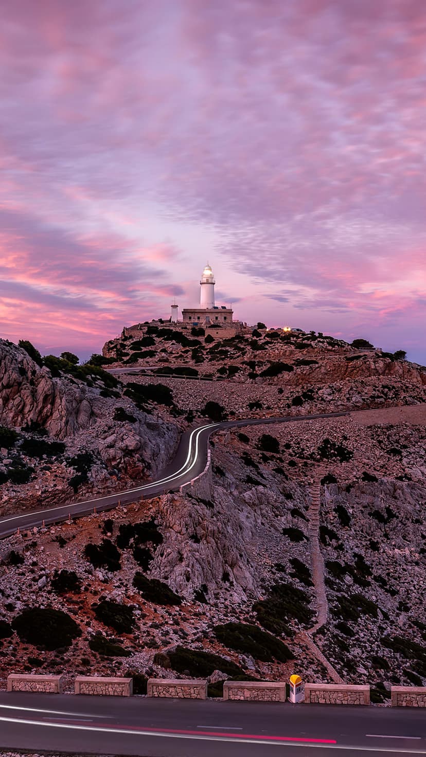 Faro de Formentor lighthouse atop a rocky mountain with winding car light trails under a pink and purple dusk sky.