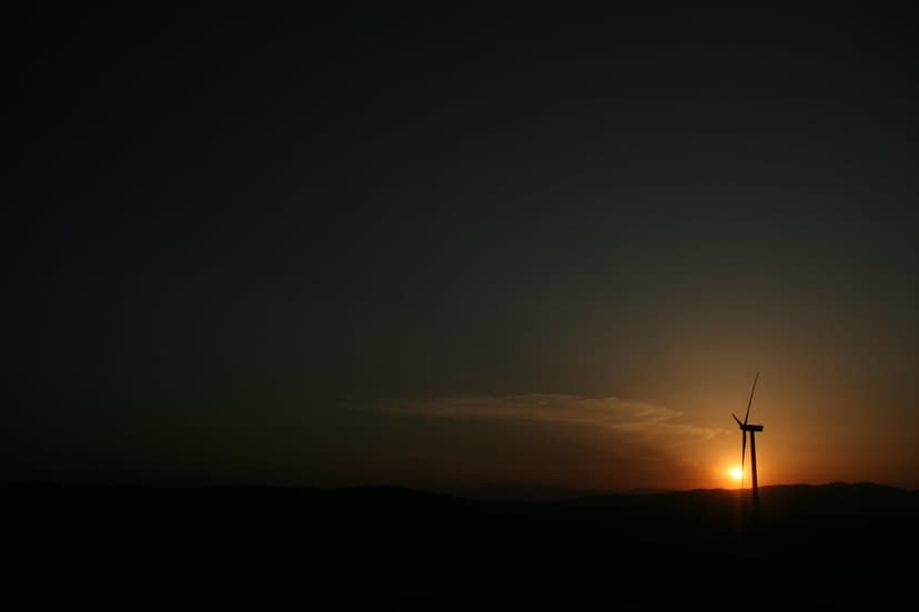 Wind turbine silhouette stands against a glowing orange sunset sky with faint clouds over a dark rural horizon.