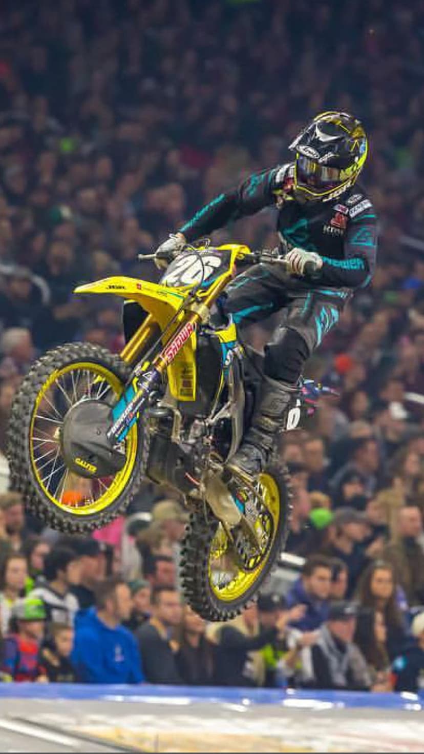 Motocross rider on a yellow motorcycle performs a mid-air stunt inside a stadium with blurred spectators behind.