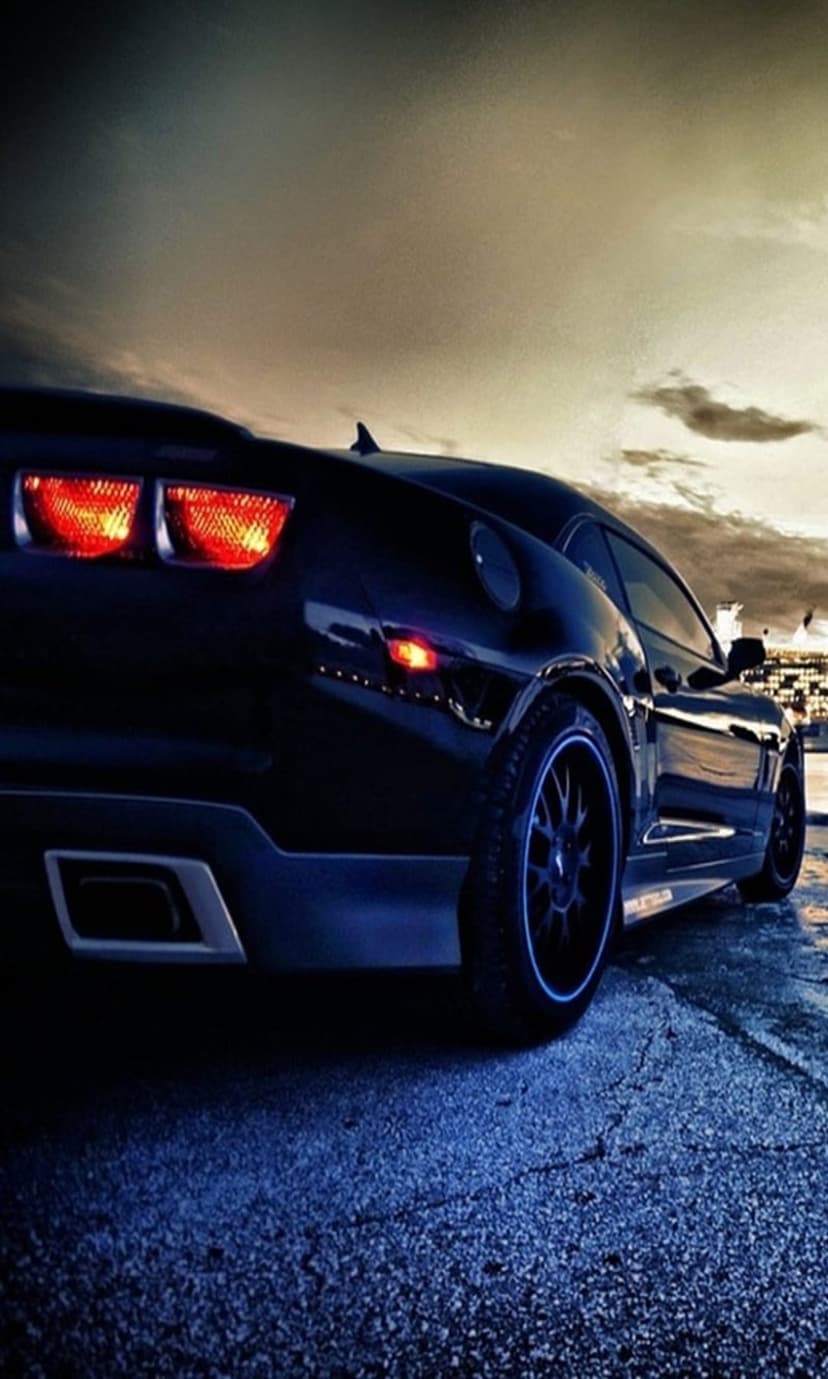Black Chevrolet Camaro rear with glowing red tail lights under a moody dusk sky on textured asphalt ground.